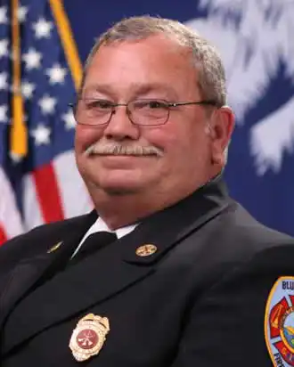 A man in a firefighter’s dress uniform with badges smiles in front of an American flag and a blue backdrop with an emblem.