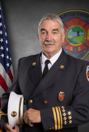 A smiling male fire chief in full dress uniform stands holding his hat. An American flag and a fire department insignia are visible in the background.