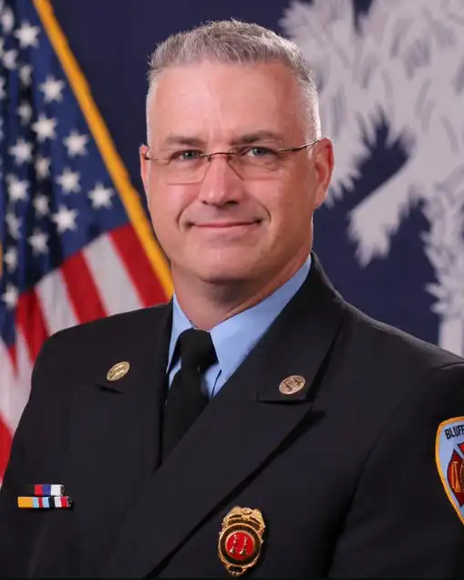 A man in a dress uniform with a badge and pins stands in front of the American flag and a blue flag with a white palmetto tree. He has short gray hair, glasses, and is smiling slightly.