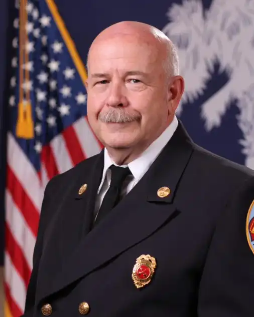 A uniformed fire chief with a bald head and mustache poses in front of an American flag and another flag with a white tree emblem.