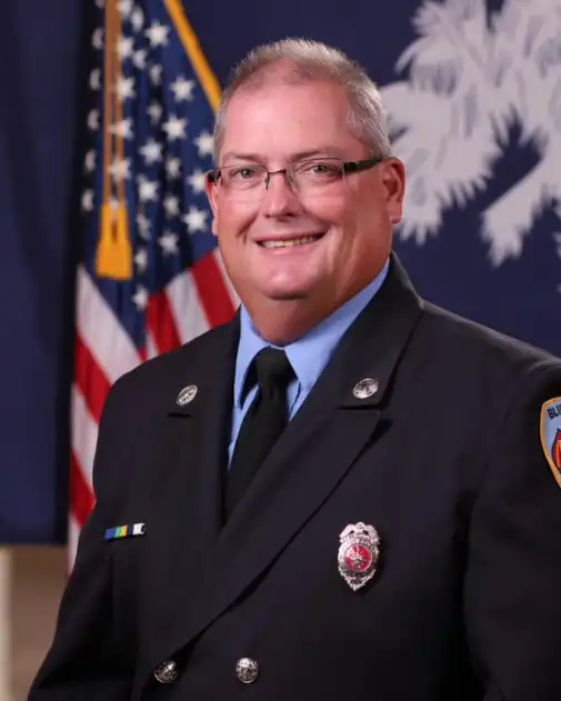 A smiling man in a fire department dress uniform stands in front of the American flag and another flag with a white tree emblem.