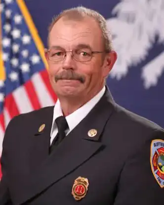 A man in a formal fire department uniform stands in front of an American flag and another flag, wearing glasses and a badge, with a mustache and short hair.