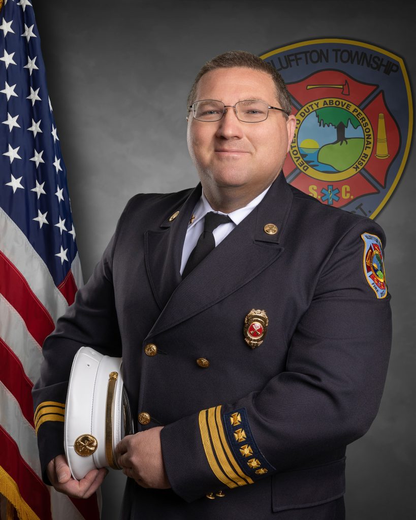 A man in a formal fire department uniform stands in front of an American flag and a Bluffton Township Fire District emblem, holding a white hat and smiling at the camera.