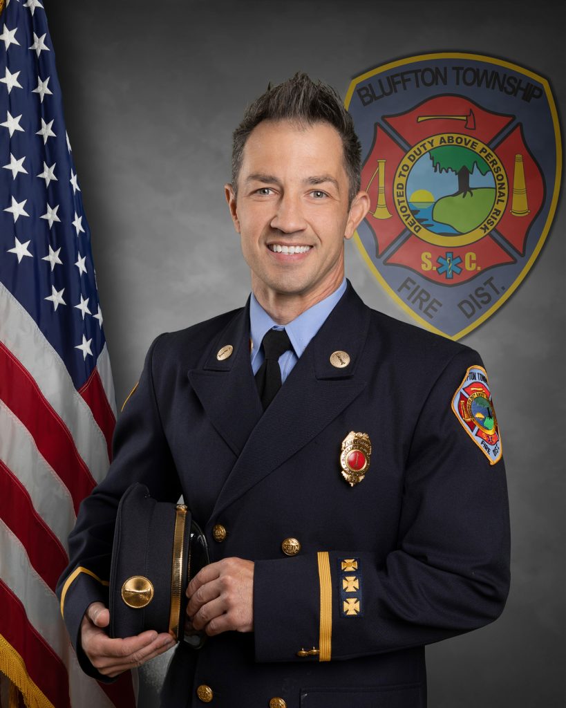 A smiling firefighter in a dress uniform holds a cap, standing beside an American flag, with the Bluffton Township Fire District badge displayed in the background.
