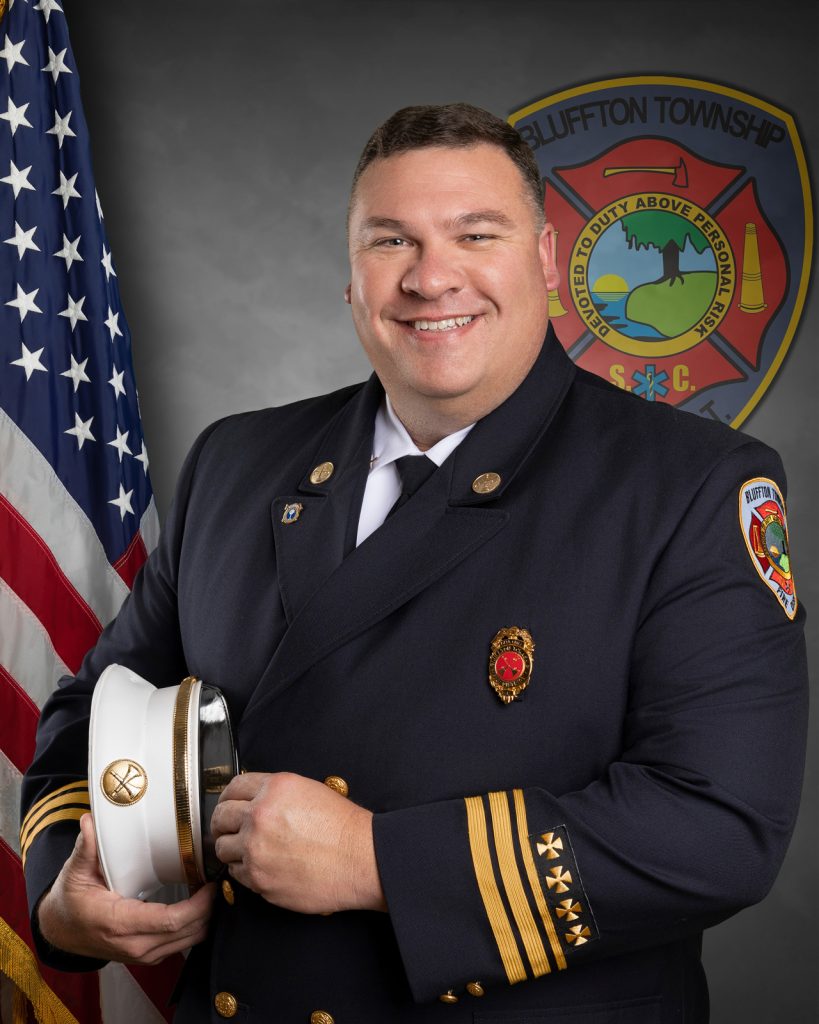 A smiling firefighter in full dress uniform holds a white hat in front of an American flag and a Bluffton Township Fire Department emblem.