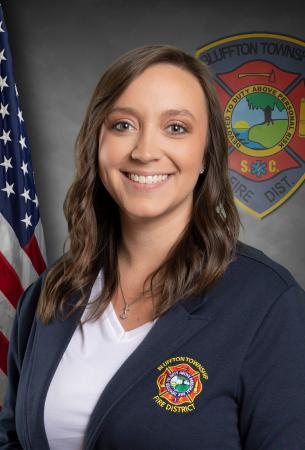 A woman with long brown hair smiles, wearing a navy Bluffton Township Fire District jacket over a white shirt. An American flag and the fire district logo are in the background.
