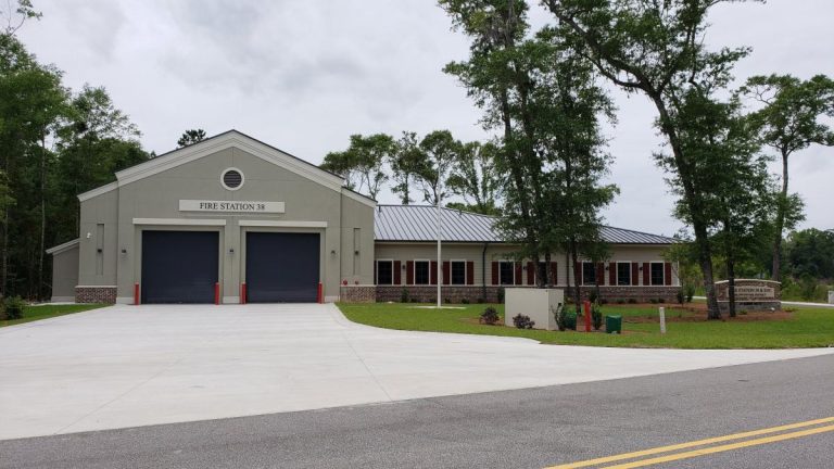 A modern fire station with two large garage bays, labeled "FIRE STATION 38," sits surrounded by grass, trees, and a driveway leading to the main road under a cloudy sky.