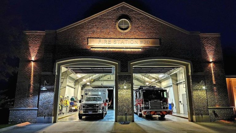 A brick fire station at night, brightly lit with two large garage doors open. Inside, a fire truck and an emergency vehicle are visible, with equipment and personnel standing nearby.