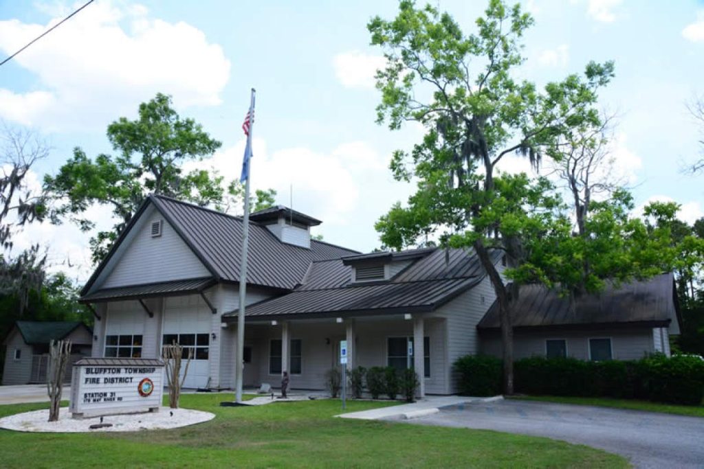 A gray building with a metal roof and porch serves as the Bluffton Township Fire District station. A flagpole and sign stand outside, surrounded by green grass and tall trees under a partly cloudy sky.