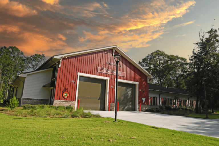 A red fire station with large garage doors and a sign reading “Station 31,” surrounded by green grass and trees, under a dramatic orange and blue sunset sky.