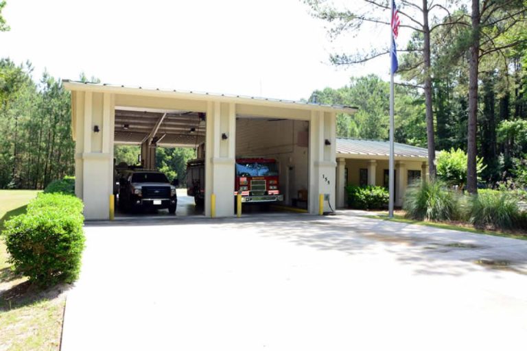 A small fire station with two garage bays open, showing a fire truck and another vehicle inside. The building is surrounded by trees, shrubs, and a neatly landscaped lawn under a clear sky.