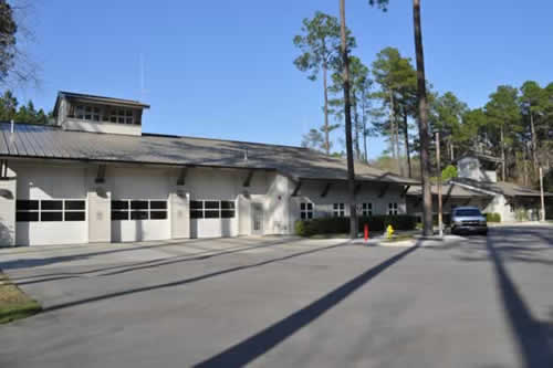 A modern fire station building with multiple garage bays and a parked pickup truck outside, surrounded by tall pine trees under a clear blue sky.