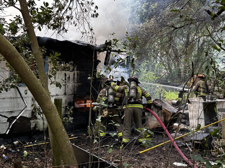 Three firefighters in gear spray water on a burning, heavily damaged house surrounded by trees and debris. Smoke rises in the background as the fire is being contained in a wooded area.