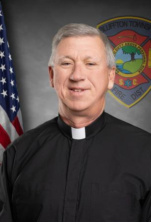 A man wearing a black clerical shirt with a white collar stands smiling in front of an American flag and a fire department emblem.