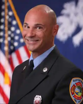 A smiling firefighter in a formal uniform stands in front of an American flag and another official flag, with an embroidered department patch visible on his sleeve.