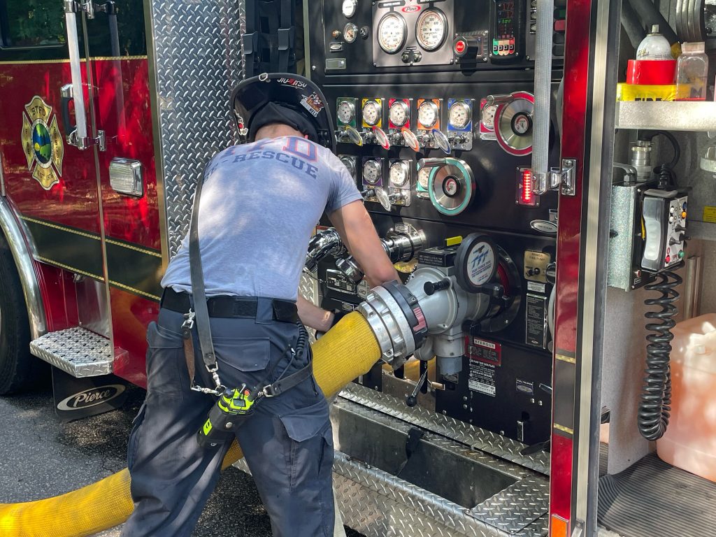 A firefighter in uniform connects a large yellow hose to a fire truck and checks gauges on the control panel during an emergency response.