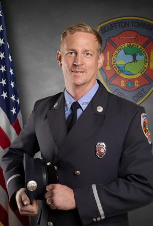 A firefighter in formal uniform holds his hat and smiles. An American flag and a Bluffton Township Fire District emblem are visible in the background.