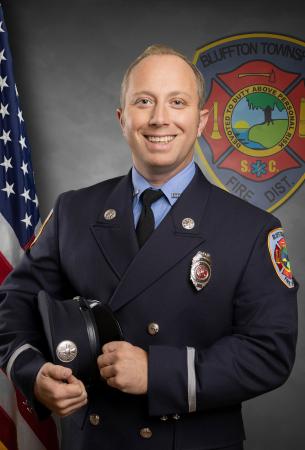A smiling firefighter in dress uniform holds a hat in front of an American flag and the Bluffton Township Fire District emblem.