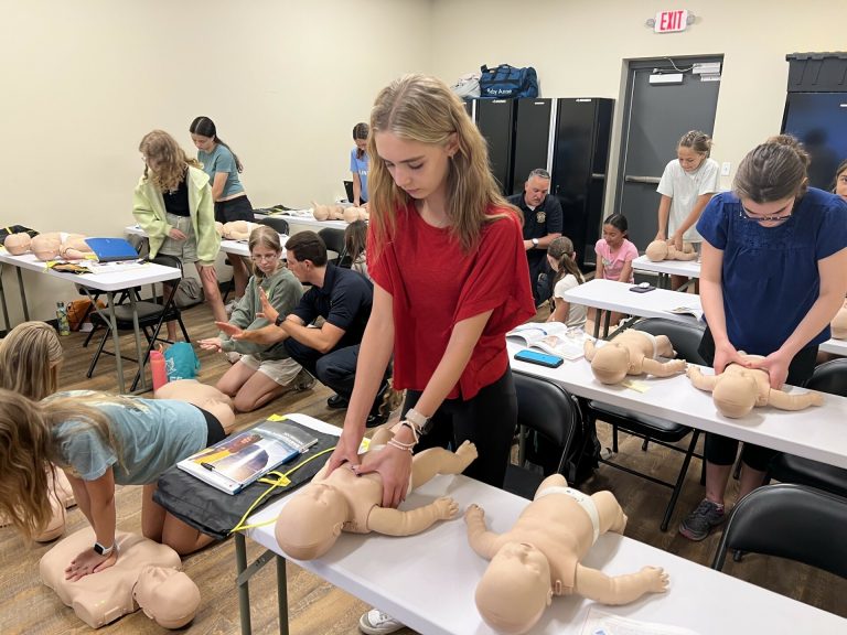 Several students practice infant CPR on training mannequins in a classroom setting, with an instructor observing. The students are focused, using their hands to perform CPR compressions.