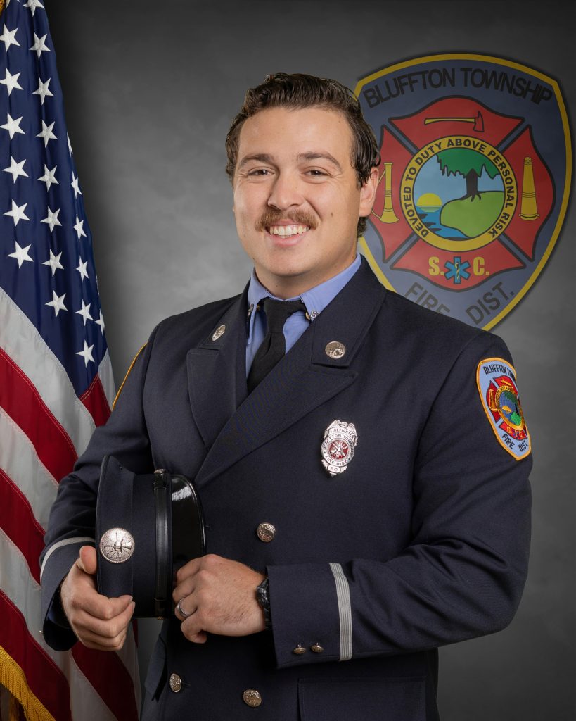 A firefighter in formal uniform smiles while holding a hat. He stands in front of an American flag and a Bluffton Township Fire District emblem on a gray background.