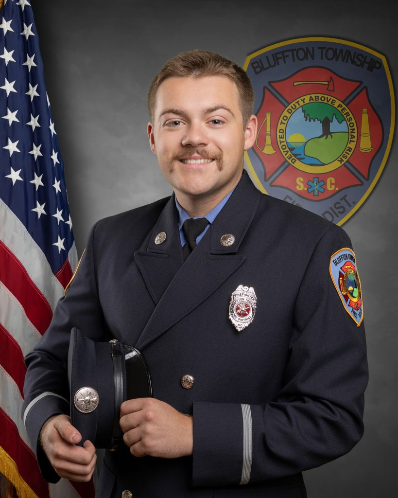 A firefighter in formal uniform poses holding his hat, standing in front of an American flag and the Bluffton Township Fire District emblem. He has short hair and a mustache, and is smiling at the camera.