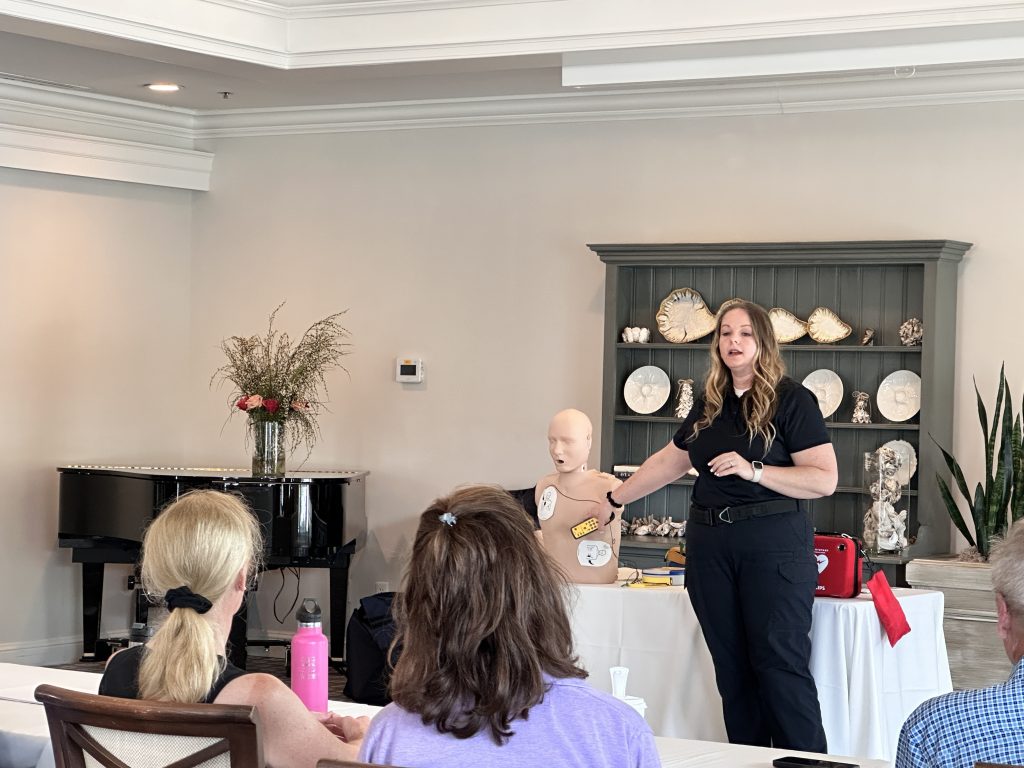 A woman demonstrates CPR on a medical mannequin in a classroom, addressing seated participants. A piano, flowers, and shelves with decorative plates are visible in the background.