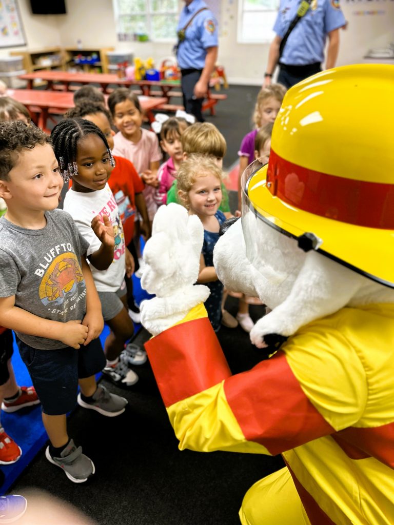 A person in a firefighter mascot costume greets a group of smiling young children in a classroom, while two uniformed officers stand in the background.