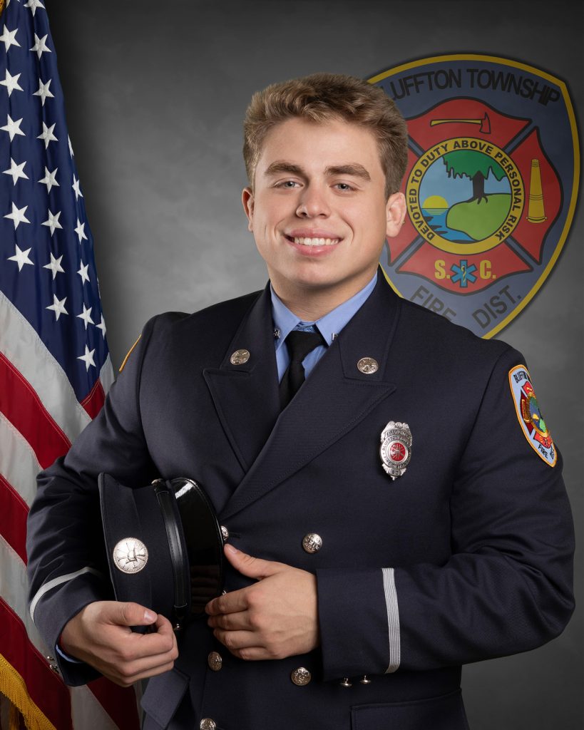 A young firefighter in formal uniform stands smiling, holding his hat. An American flag and a Bluffton Township Fire District emblem appear in the background.