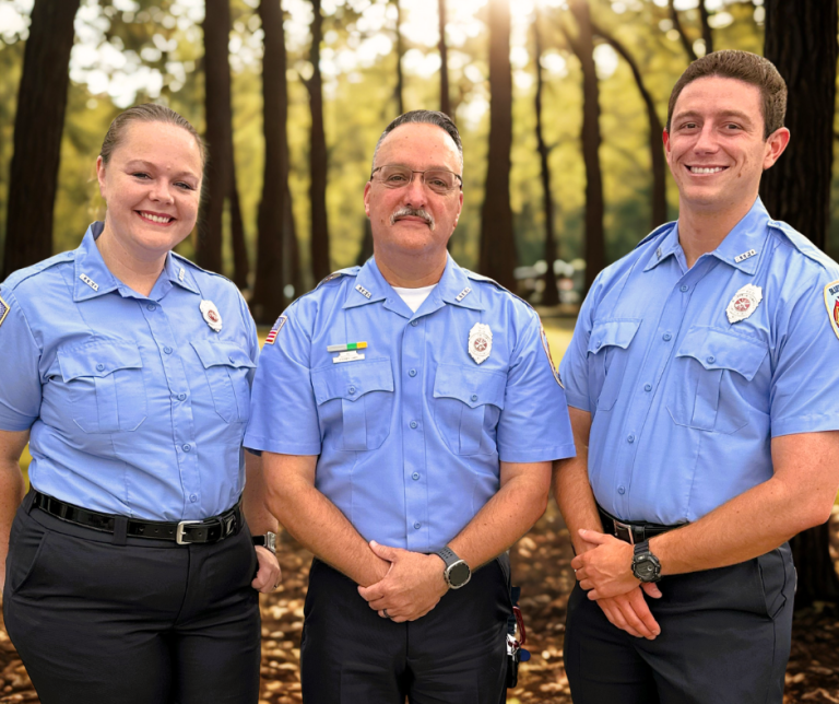 Three uniformed firefighters stand outdoors in a wooded area, smiling at the camera. Sunlight filters through the trees in the background.