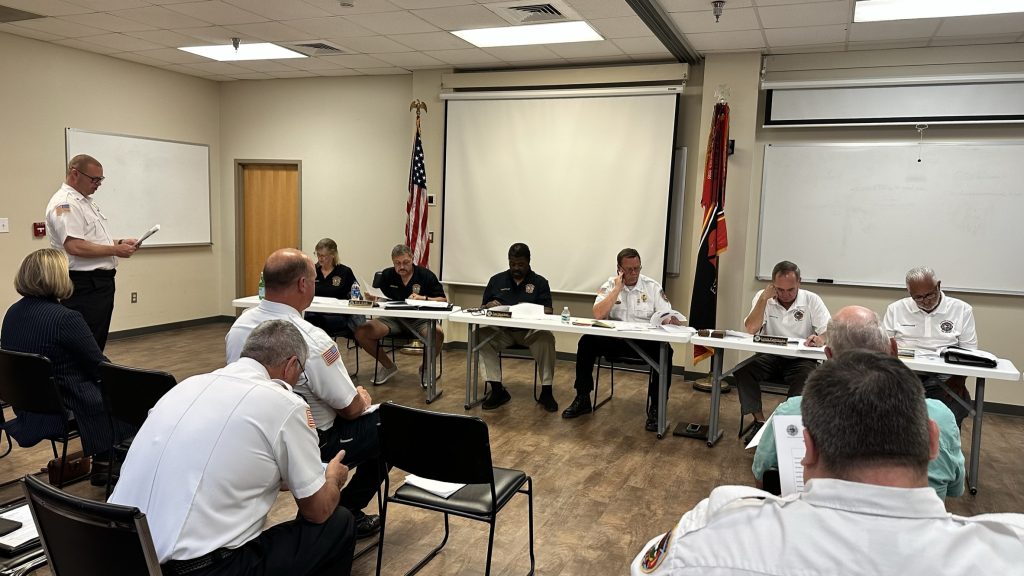 A group of people in uniform sit at a long table in a meeting room, with others seated in the audience. An American flag and state flag are in the background, and one man stands reading from a paper.
