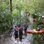 Four emergency responders stand in knee-deep water surrounded by dense greenery, with two empty orange kayaks nearby. The group appears to be conducting a rescue or search operation in a flooded forest area.