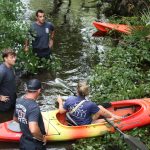 Four people, three standing in shallow, leafy water and one sitting in an orange kayak, appear to be discussing something. The standing individuals wear shirts labeled "FIRE DEPT" and are surrounded by dense greenery.