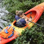 A person in a bright orange kayak paddles through green water plants, carrying a blue and orange life jacket behind them. The person wears a navy shirt with a graphic on the back.