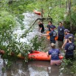 Several people in navy shirts stand in shallow, wooded water guiding an orange kayak through branches and greenery, with more people and another kayak in the background.