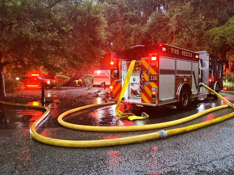 A fire truck with hoses extended is parked on a wet street at dusk, with emergency lights flashing. Firefighters and an ambulance are visible in the background near trees.