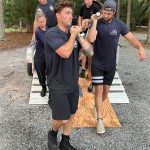 Five people in athletic clothing work together to carry one person on a wooden board over a narrow bridge outdoors, surrounded by trees and gravel. Teamwork and concentration are evident in their expressions and posture.