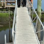 Three emergency responders assist a person on a stretcher down a sloped metal ramp from a wooden dock, with marsh grass and water visible in the background.