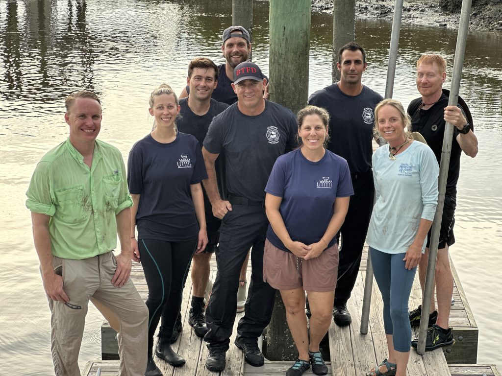 A group of nine people stands on a wooden dock by the water, smiling at the camera. They are casually dressed, some in T-shirts with logos, and appear to be part of an outdoor or marine activity team.