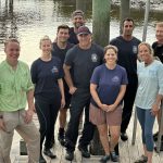 Nine people stand and smile on a wooden dock by the water, dressed in casual and work clothing, some in uniforms with a logo. The group appears relaxed and friendly, with boats or pilings visible in the background.