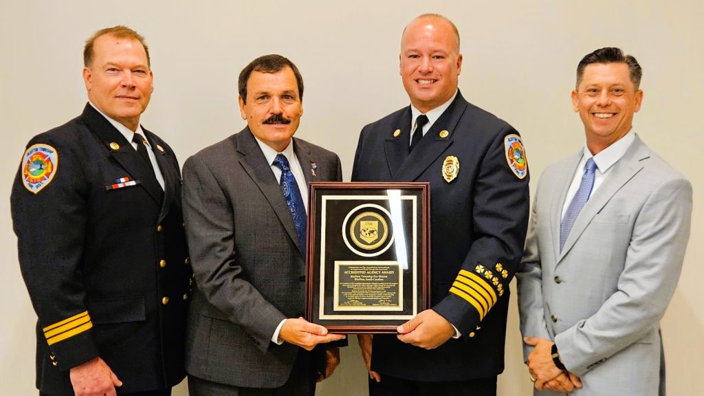 Four men in formal attire stand together smiling; one in a fire chief uniform holds a framed award plaque, flanked by two men in uniforms and one man in a suit, against a plain light-colored background.