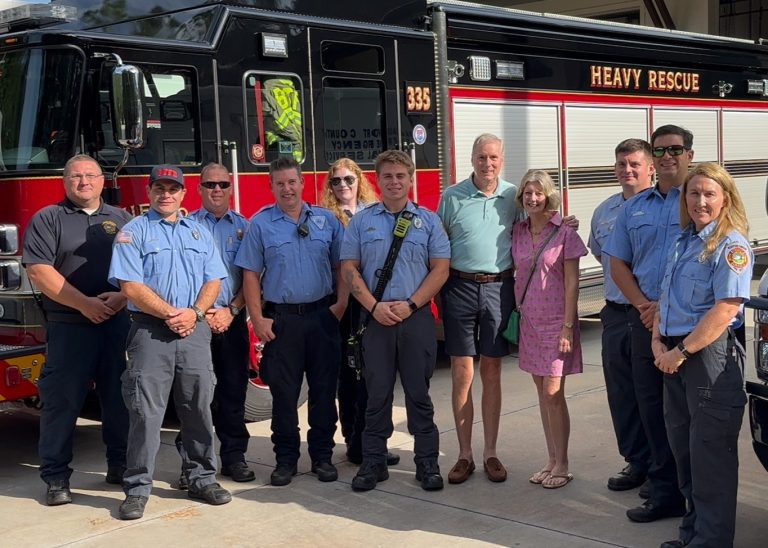 A group of firefighters in uniform and two civilians stand together in front of a Heavy Rescue fire truck on a sunny day, smiling for the photo.
