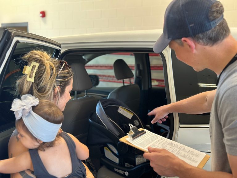 A woman holding a young child stands by an open car door while a man with a clipboard points at a car seat inside the vehicle, possibly giving car seat safety instructions.