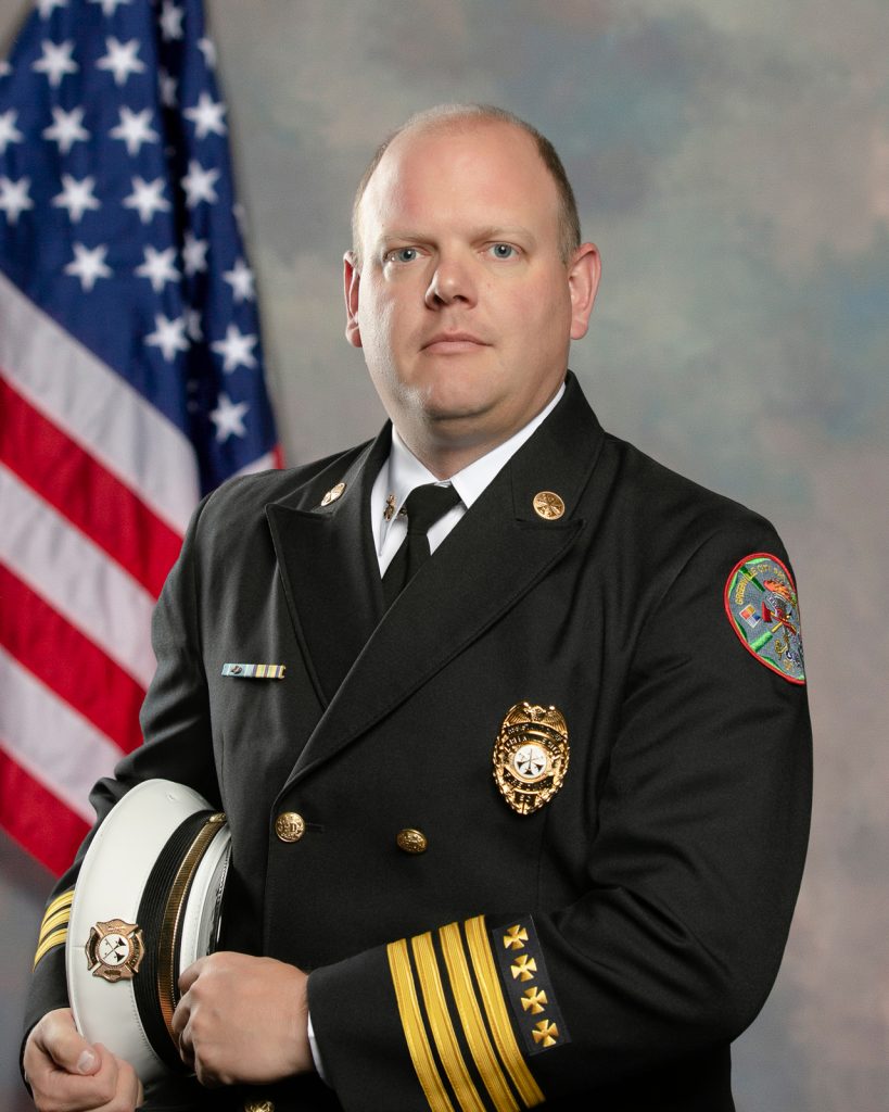 A uniformed fire chief stands in front of an American flag, holding a white service cap. He wears a dark dress uniform with badges, medals, and gold-striped sleeves, and looks directly at the camera with a serious expression.