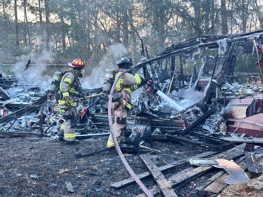 Two firefighters in full gear spray water on the smoking remains of a burnt-down wooden structure, surrounded by debris and charred wood, with trees visible in the background.