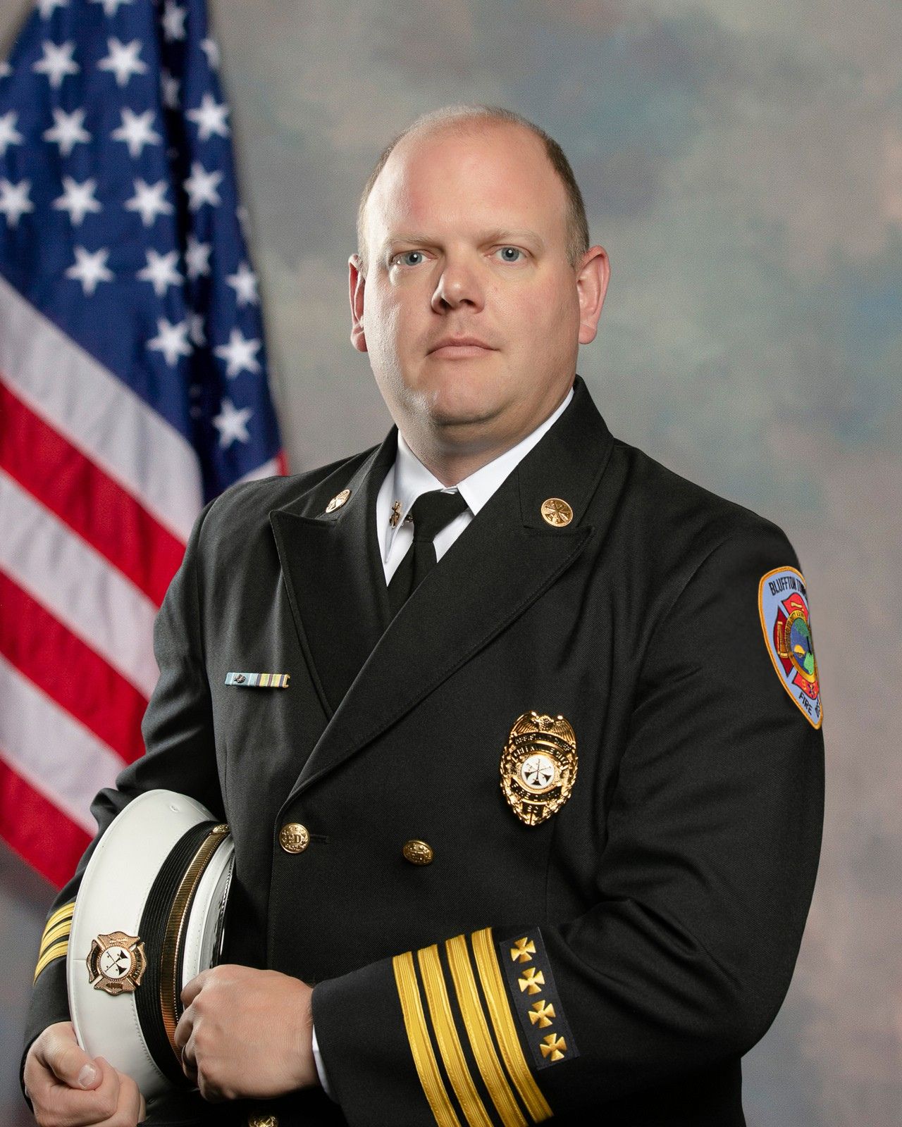A uniformed fire department officer stands holding a white hat, wearing a black jacket with gold insignia and a badge. An American flag is in the background. The officer has a serious expression and short hair.