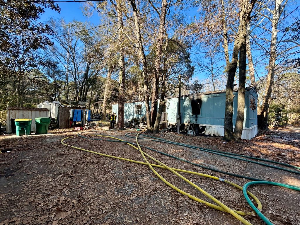 A blue mobile home surrounded by trees, with several long yellow and green hoses spread across the leaf-covered ground. Green trash bins and a blue dumpster are visible nearby under a clear blue sky.