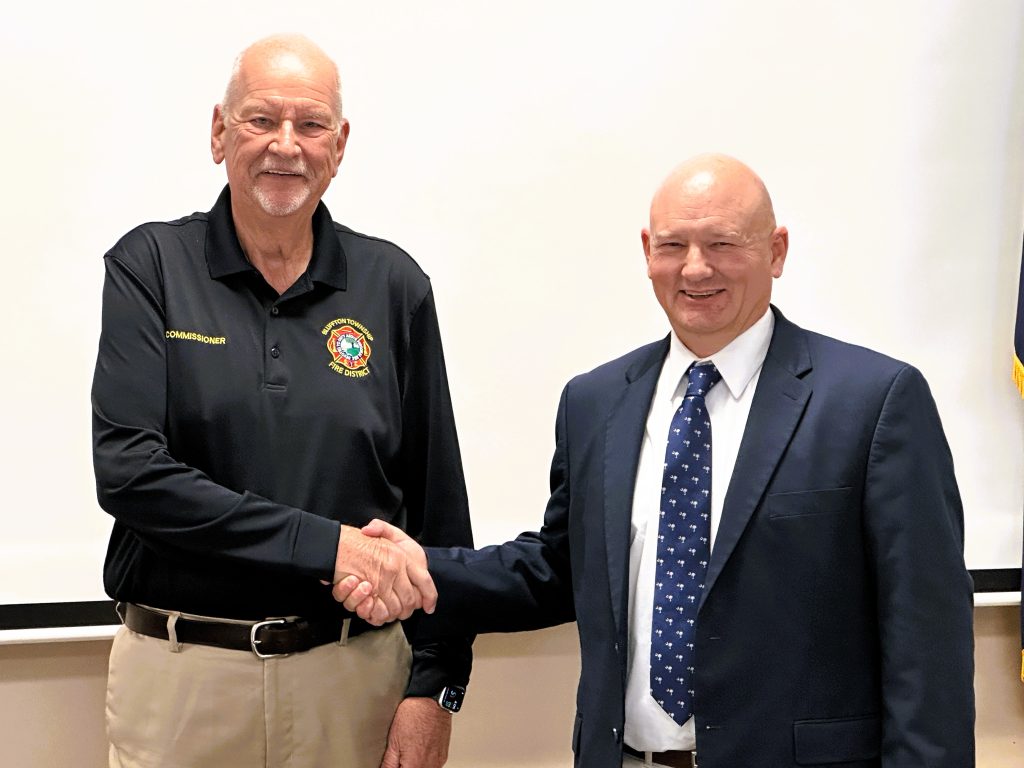Bluffton Township Fire District Board Chairman Bill Rickett (left), wearing a black department polo, shakes hands with newly appointed board member Glenn Williams (right), who is dressed in a dark suit and tie. The two stand side by side in front of a neutral background following Williams’ swearing-in ceremony on Tuesday, marking his official appointment to the Fire Commission Board.