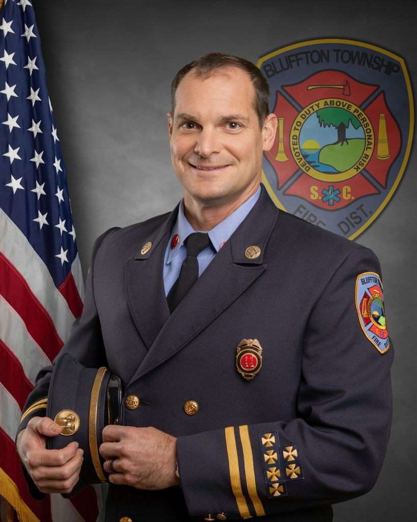 A firefighter in a formal uniform stands holding his hat, smiling in front of a U.S. flag. The Bluffton Township Fire District emblem is displayed on the wall behind him.