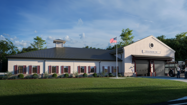 A modern, single-story fire station with beige siding, red shutters, and a metal roof. An American flag flies out front, and a fire truck is partially visible at the garage entrance labeled "Station 50.