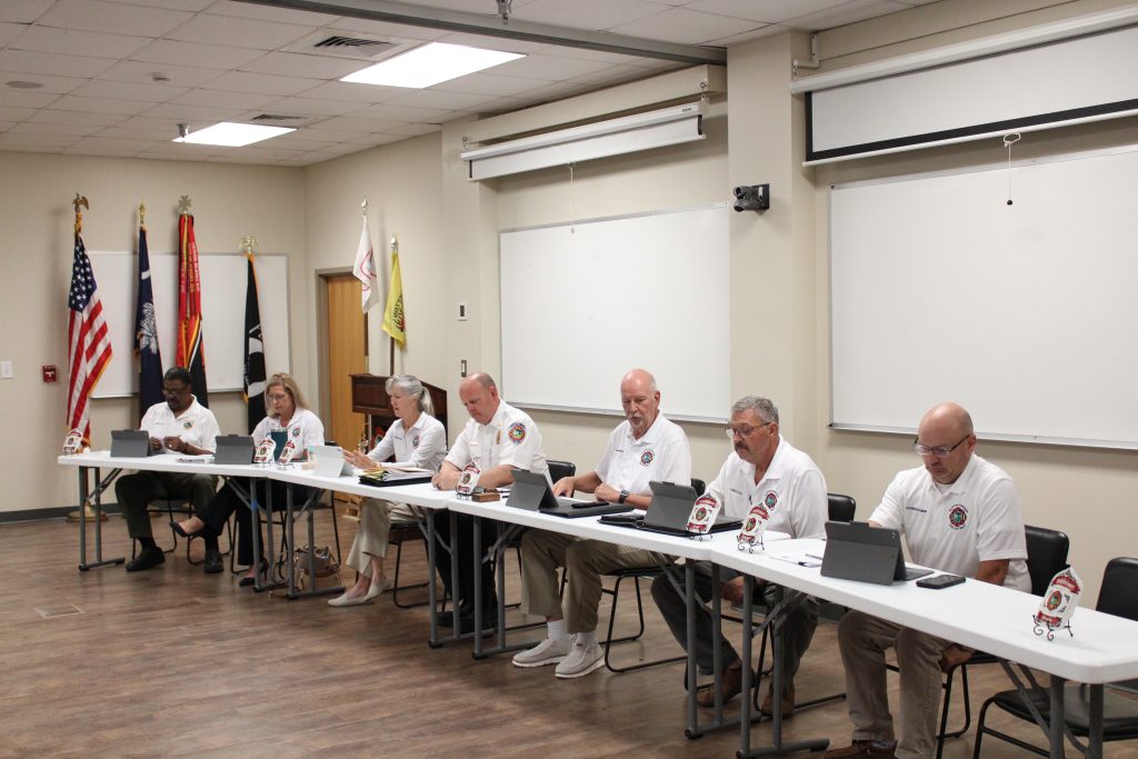 Seven people in matching white polo shirts sit at a long table with laptops and papers in a meeting room, with flags and whiteboards in the background.